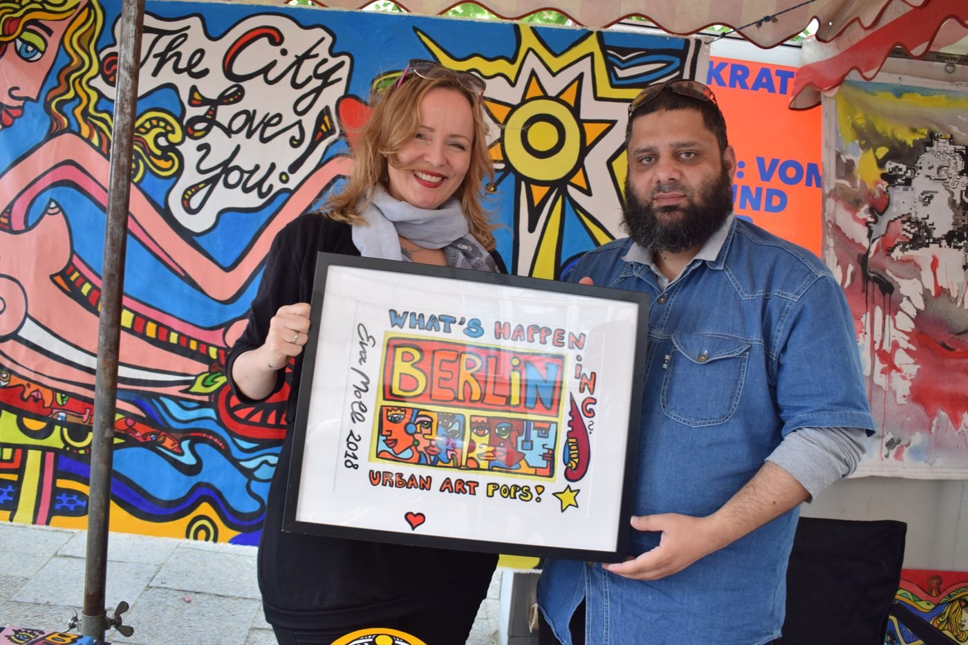 Dr. Ahmed Ejaz with an artist holding a framed What&rsquo;s Happening Berlin urban art piece