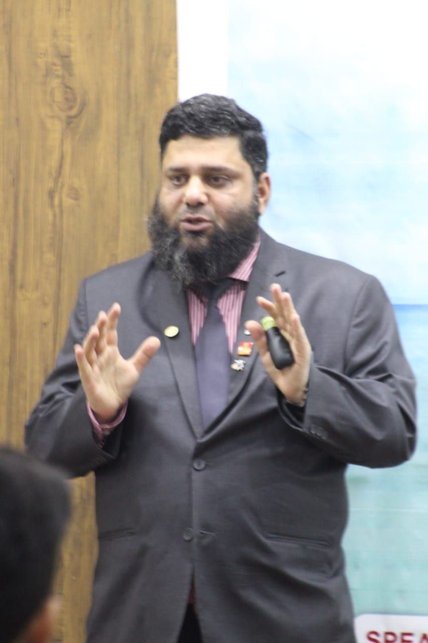 Dr. Ahmed Ejaz in a dark suit with Rotary pins, speaking with both hands raised